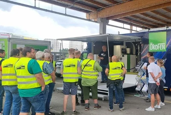 Gruppe Heizungsbauer mit neongelben Westen vor Roadshow-Container mit Heiztechnik von ÖkoFen.