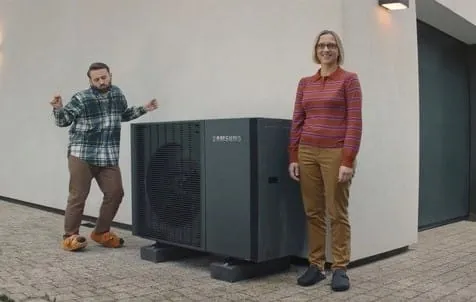 Mann und Frau stehen vor ihrem Haus. Zwischen ihnen eine eingebaute W&auml;rmepumpen-Au&szlig;eneinheit von Samsung. Mann tanzt vor Freude in albernen Tier-Hauspuschen.