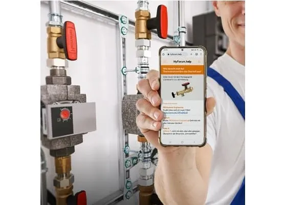 Handwerker zeigt Smartphone in die Kamera mit Bildschirmansicht HyForum. Im Hintergrund Steigstr&auml;nge Heizung im Heizungsraum.