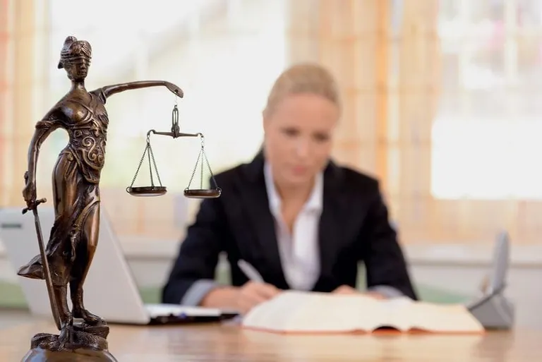 Bronzefigur von Justizia auf Schreibtisch, im Hintergrund verschwommen eine Frau in Bussinesskleidung am Schreibtisch.