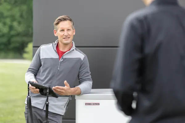 SHK Handwerker lehnt an W&auml;rmepumpe vor einem Haus. Im Vordergrund verschwommen ein Kunde.