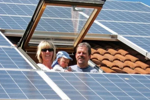 Mutter und Vater mit Kleinkind in der Mitte schauen aus Dachfenster eines PV-Daches und l&auml;cheln.