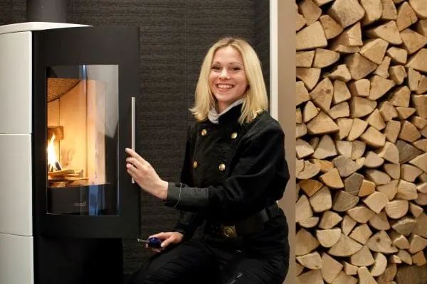 Schornsteinfegerin vor modernen Holzofen. Sie &ouml;ffnet die Ofent&uuml;r. Im Hintergrund gestapelte Holzscheite