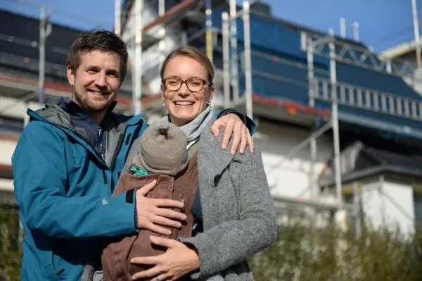 Junges Paar vor Einfamilienhaus mit Ger&uuml;st. Die Frau tr&auml;gt ein Baby im Tragetuch vor dem Bauch. Der Mann legt seine Hand auf den R&uuml;cken des Babys.