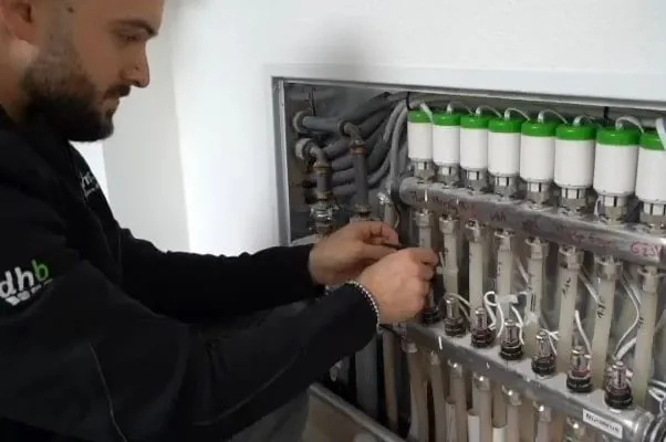 Monteur vor Stellventilen einer Fußbodenheizung im Einbauschrank. Er installiert automatische Stellventile für den digitalen hydraulischen Abgleich.