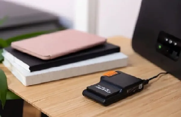 Solarwatt Manager, ein kleines schwarzes Ger&auml;t in Handygr&ouml;&szlig;e mit orangen Taster an der Seite liegt auf einer Holztischplatte neben drei aufgestapelten Notizb&uuml;chern und vor einem Router