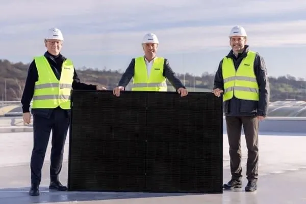 Drei Mitarbeiter mit neongelben Warnwesten und wei&szlig;en Schutzhelmen auf einem Flachdach gruppiert um ein PVT-Modul von Sunmaxx