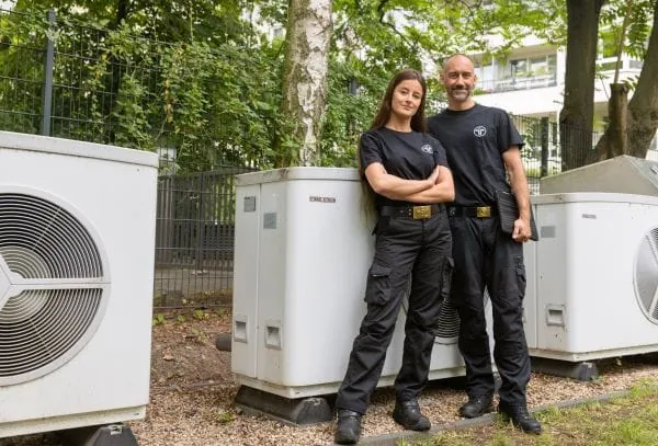 Schornsteinfegerin und Schornsteinfeger vor W&auml;rmepumpen in einem Wohngebiet.