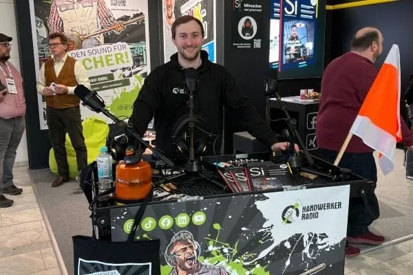 Moderator Max hinter rollendem Podcast-Studio, dahinter Messestand Si