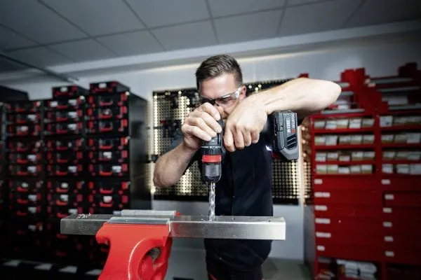 Handwerker bohrt mit Borhrmaschine in ein Blech, eingespannt im Schraubstock. Im Hintergrund gelagerte Kleinteile in Regalen.