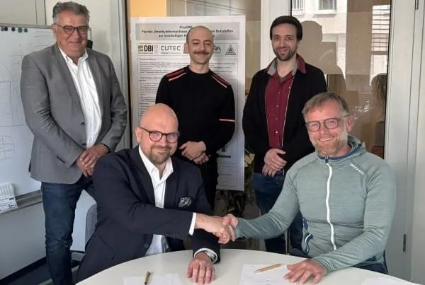 Im Vordergrund zwei Geschäfstführer sitzend am Tisch reichen sich die Hand für Vertragsabschluss. Im Hintergrund drei Mitarbeiter stehend.