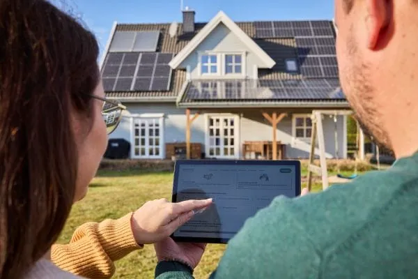Mann und Frau mit Tablet-PC und Screen PhotovoltaikCheck, in der Mitte gehalten. Im Hintergrund ein Einfamilienhaus mit PV-Anlage auf dem Dach und Vordach
