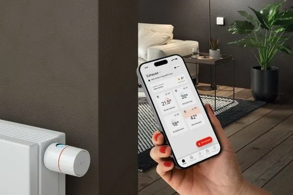 Hand h&auml;lt Smartphone und Viessmann-App auf dem Display. Im Hintergrund Heizk&ouml;rper mit Thermostatventil und Wohnzimmer