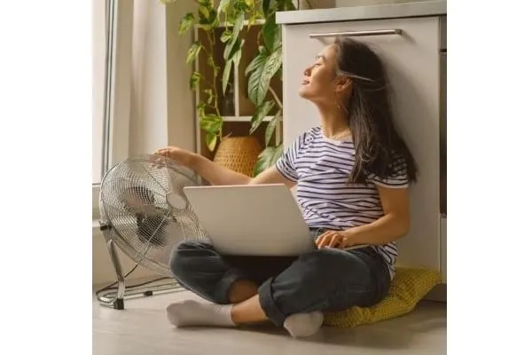 Symbolbild Prosumer: junge Frau sitzt auf dem Boden mit Laptop auf den Knien vor einem Küchenschrank. Daneben Ventilator (betrieben mit eigenen PV-Strom)