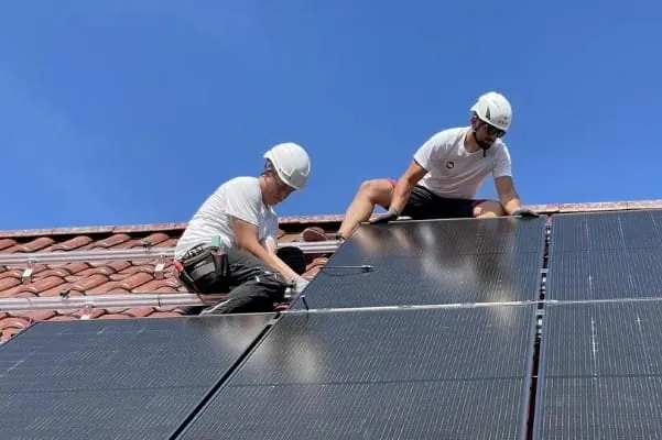 2 Installateure mit Bauhelm auf dem Dach Einfamilienhaus bei der Montage von PV-Modulen. Himmelblauer Himmel.