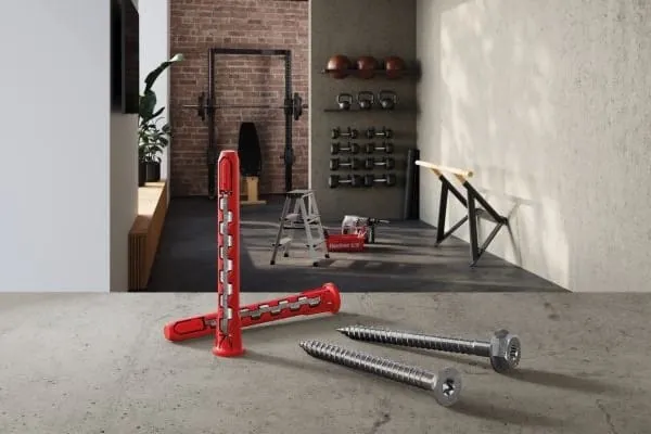 im Vordergrund zwei rote Dübel Typ HybridPower und zwei Kreuzschlitzschrauben. Im Hintergrund häusliches Fitnesstudio mit Geräten