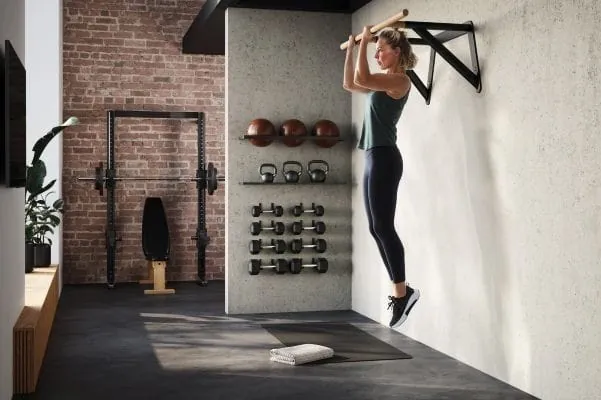 Frau macht Klimmzüge an Stange, die mit Stahlkonstruktion an der Wand in einem häuslichen Fitness-Studio befestigt ist. Im Huntergrund Kraftsportgeräte wie Hanteln und weitere Fitnessgeräte