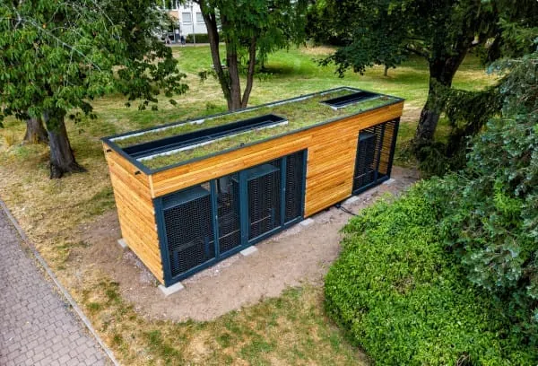 Modulare Energiezentrale f&uuml;r Wohnh&auml;user, Container mit Holzlamellen verkleidet auf einer Wiese mit B&auml;umen zwischen Wohnbauten.in