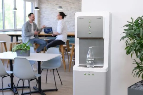 Wasserspender in einer Kantine mit halbgefüllter Glasflasche, verschwommen im Hintergrund 2 Mitarbeitende