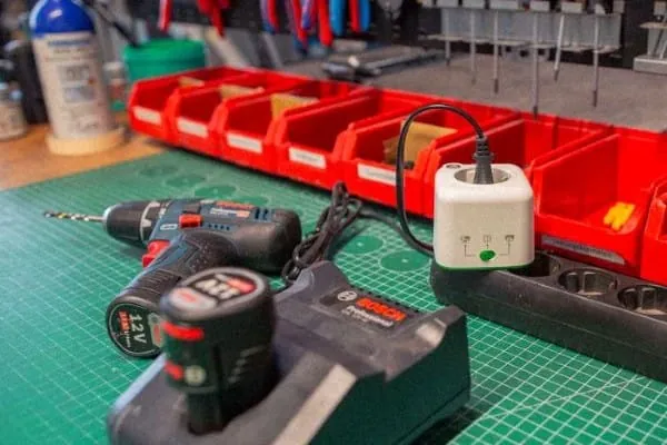 Steckdosenadapter für Akkus auf Steckdosenleiste auf Werkzeugbank. Im Vordergrund Akkubohrmaschine mit Ladestation, die mit Akku-Adapter verbunden ist