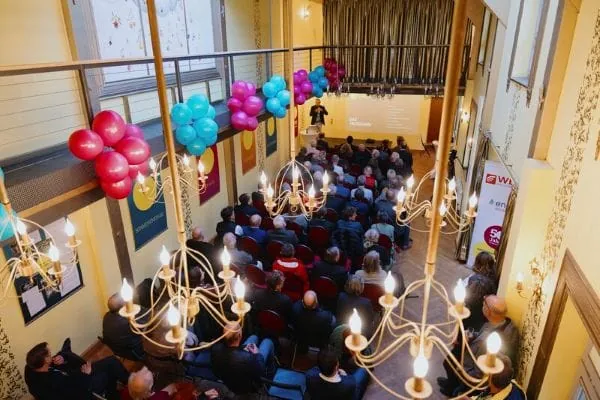 Jubil&auml;umsg&auml;ste sitzend auf Stuhlreihen h&ouml;ren den Rednern zu, Saal mit bunten Luftballons geschm&uuml;ckt