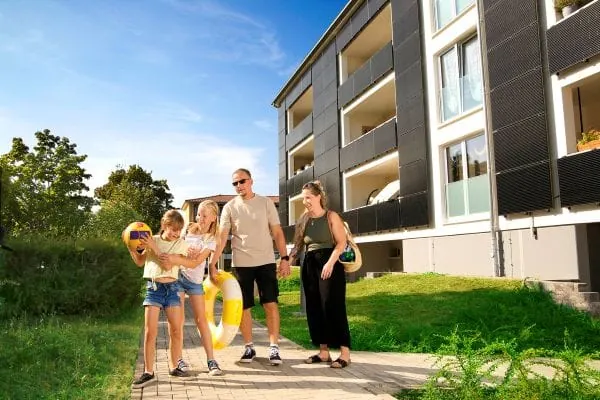 Familie mit Mutter, Vater und zwei Kindern im Grundschulalter vor Mehrfamilienhaus mit PV-Fassade. Es ist Sommer, ein Kind h&auml;lt einen Ball in den H&auml;nden, das andere einen Schwimmreifen.