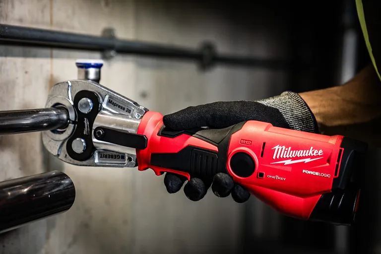 Akku-Presswerzeug in der Hand von einem Handwerker mit Arbeitshandschuh. Er verpresst damit Rohre an der Wand.