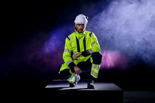 Fachkraft Energiewirtschaft mit neongelber Arbeitskleidung und Schutzhelm knieend bei einer Modeschau, düsterer Hintergrund