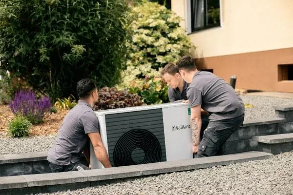 Drei Monteure tragen eine W&auml;rmepumpe von Vaillant auf Baustelle Einfamilienhaus