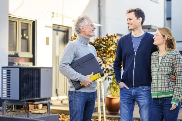 Energieberater im Kundengespr&auml;ch mit einem jungen Paar. Im Hintergrund W&auml;rmepumpe