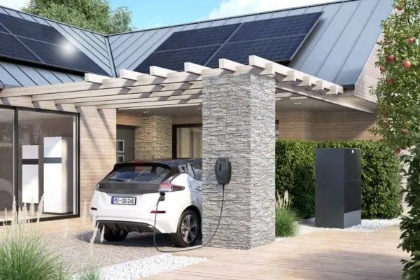 e-Auto steht unter Vordach und lädt aus der Wallbox. Im Hintergrund EFH mit PV-Dach.