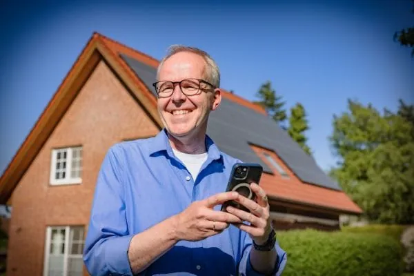 Lachender Hausbesitzer vor Eigenheim mit PV-Anlage hält ein Handy in der Hand.