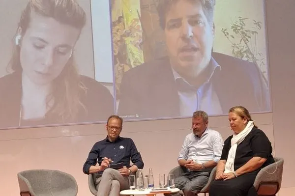 3 Personen auf Podiumsdiskussion. Im Hintergrund Screen mit zwei zugeschalteten Teilnehmern